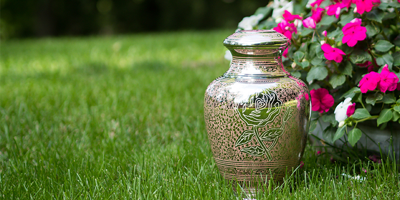 A mirror-like metal urn sitting in grass with a bush of flowers to it's right.